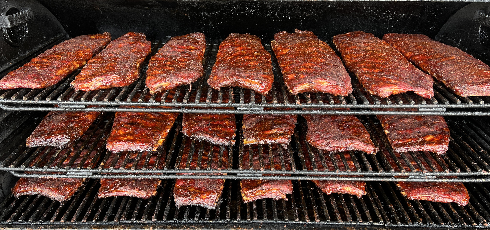 Racks of ribs with dark bark cooking on a multi-tier smoker
